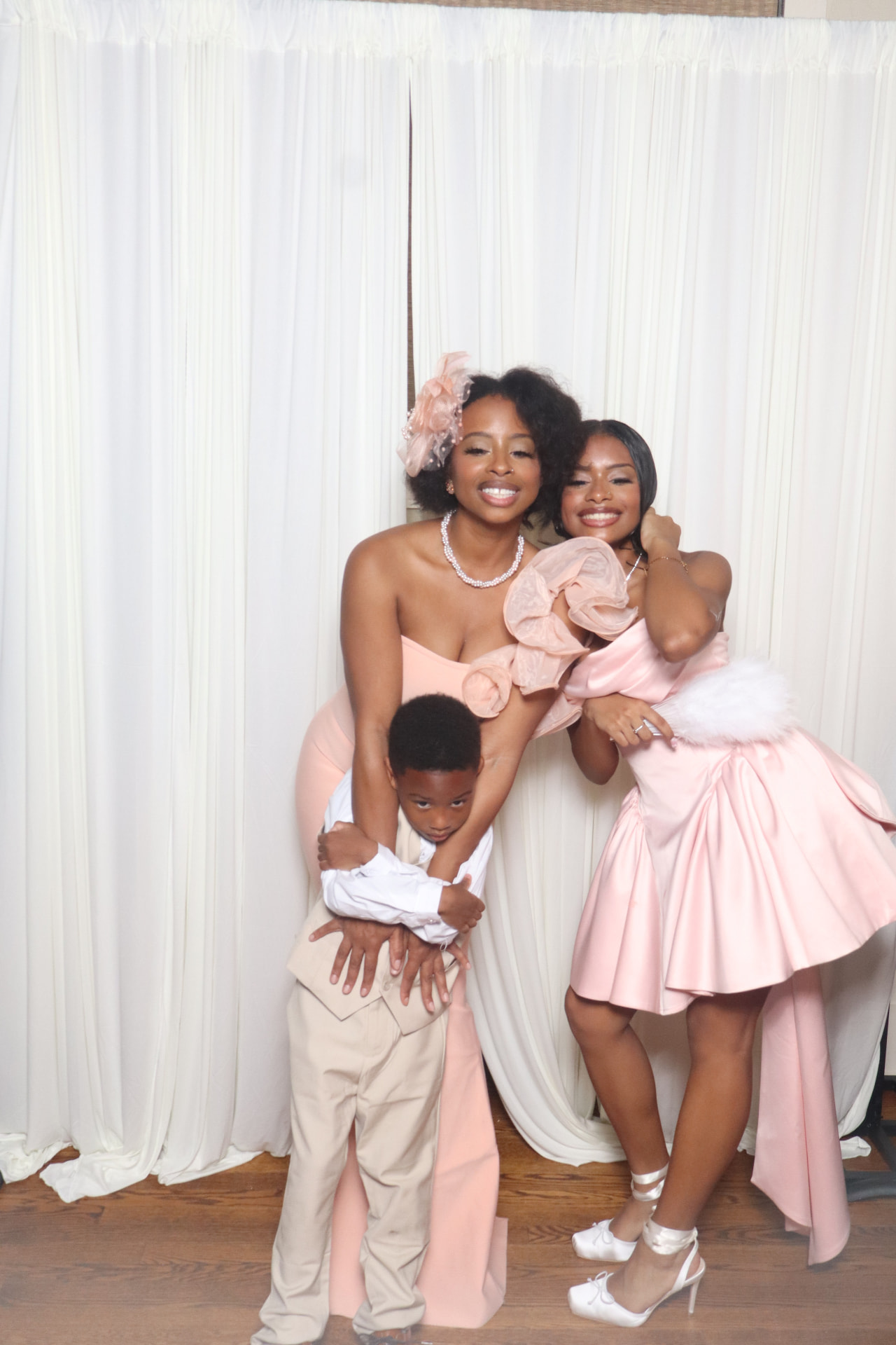 Two guests in pink and a young boy hugging in front of an ivory drape